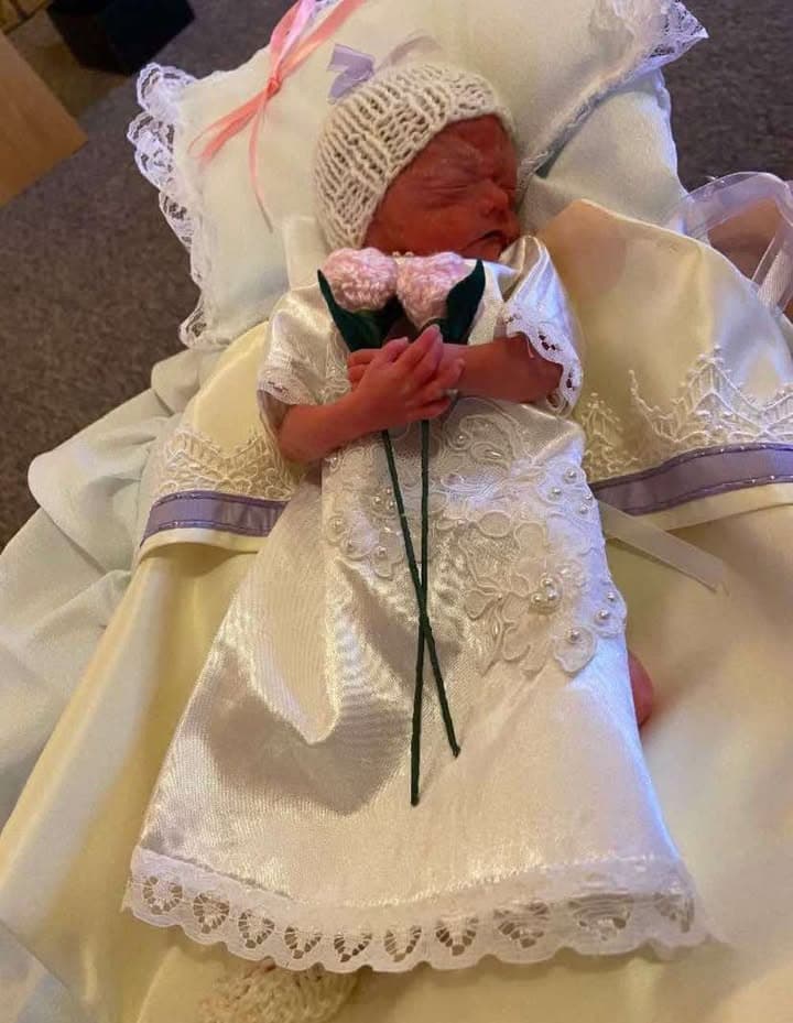 Baby Everley in a white lace gown and knitted cap, holding a small bouquet of pink knitted flowers