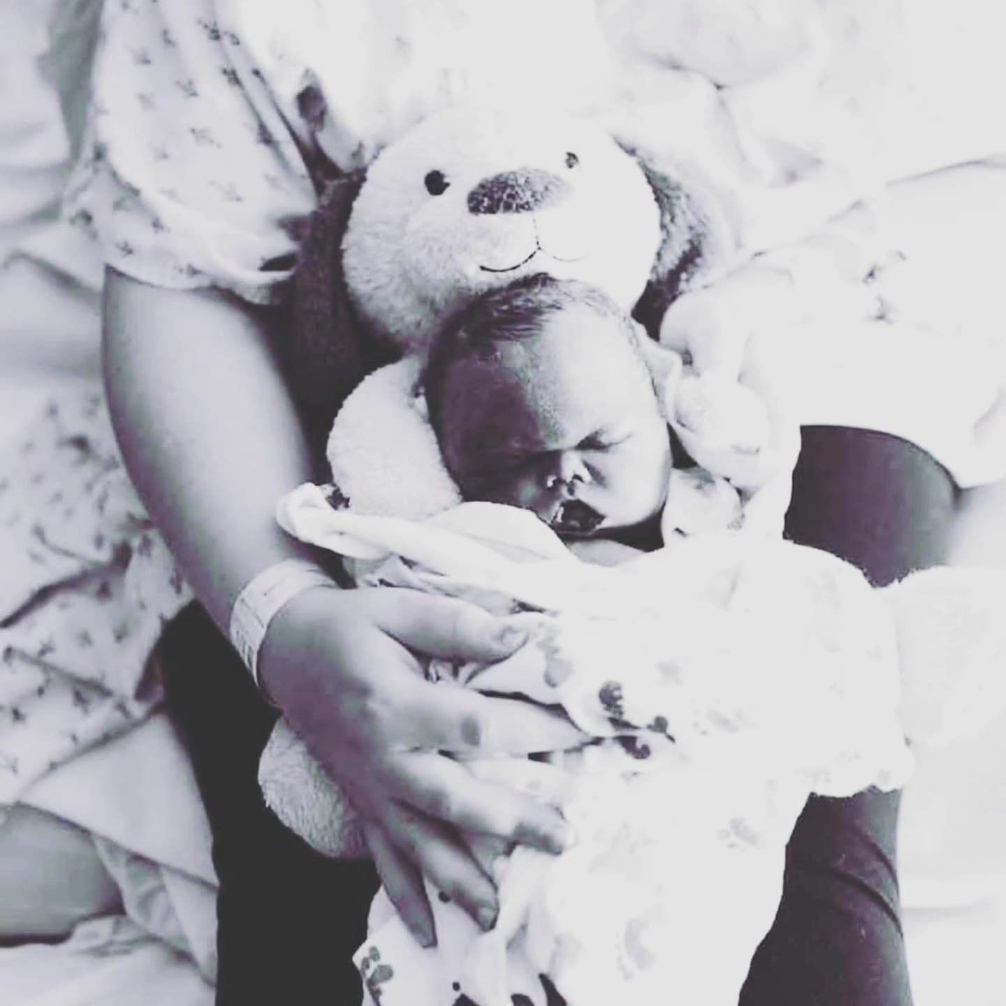 Black-and-white photo of baby Kacen cradled in his mother's arms beside a teddy bear