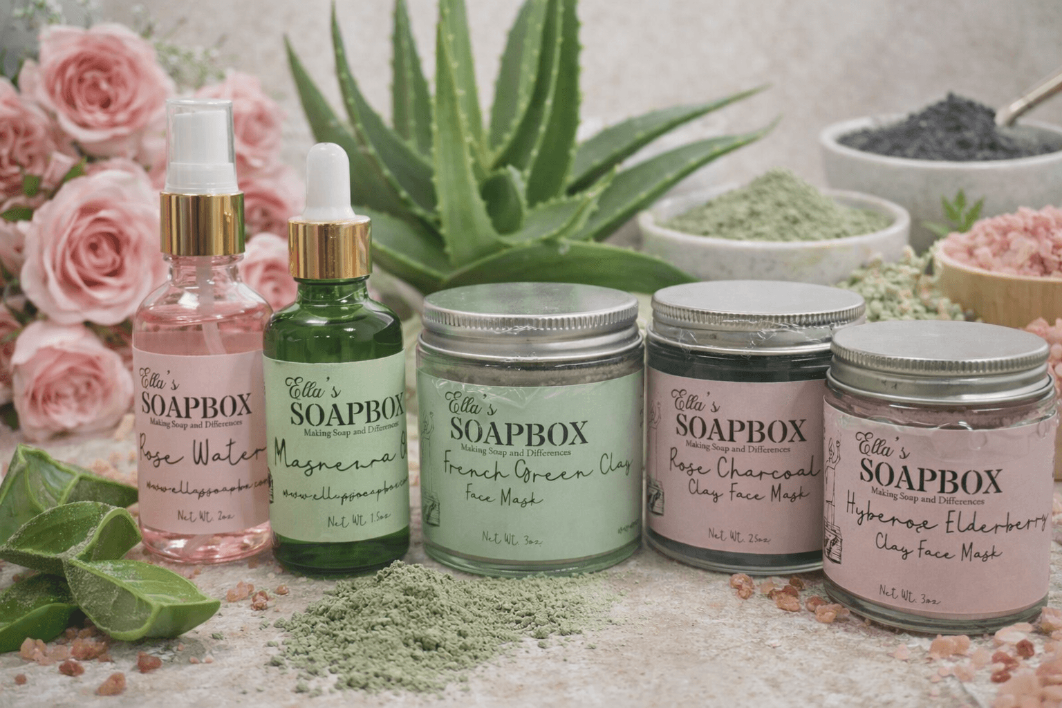 Ella's Soapbox clay face mask jars (French Green Clay, Rose Charcoal and Hibiscus Elderberry) beside bottles of Rose Water and Aloe Vera Oil, with roses, aloe and bowls of clay powder