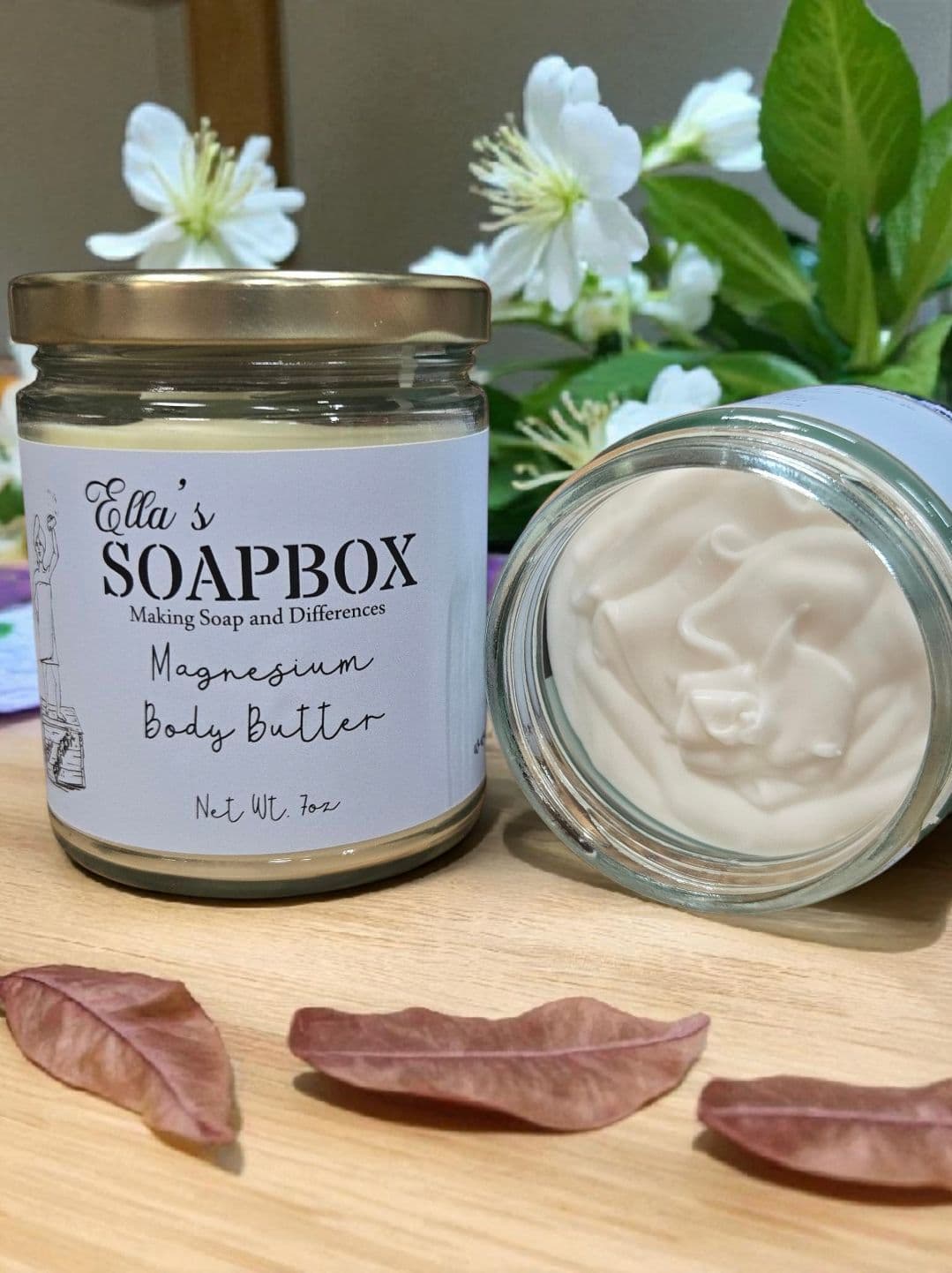 Two jars of Ella's Soapbox Magnesium Body Butter on a wooden table, one open to show the whipped cream-colored butter, with white blossoms behind