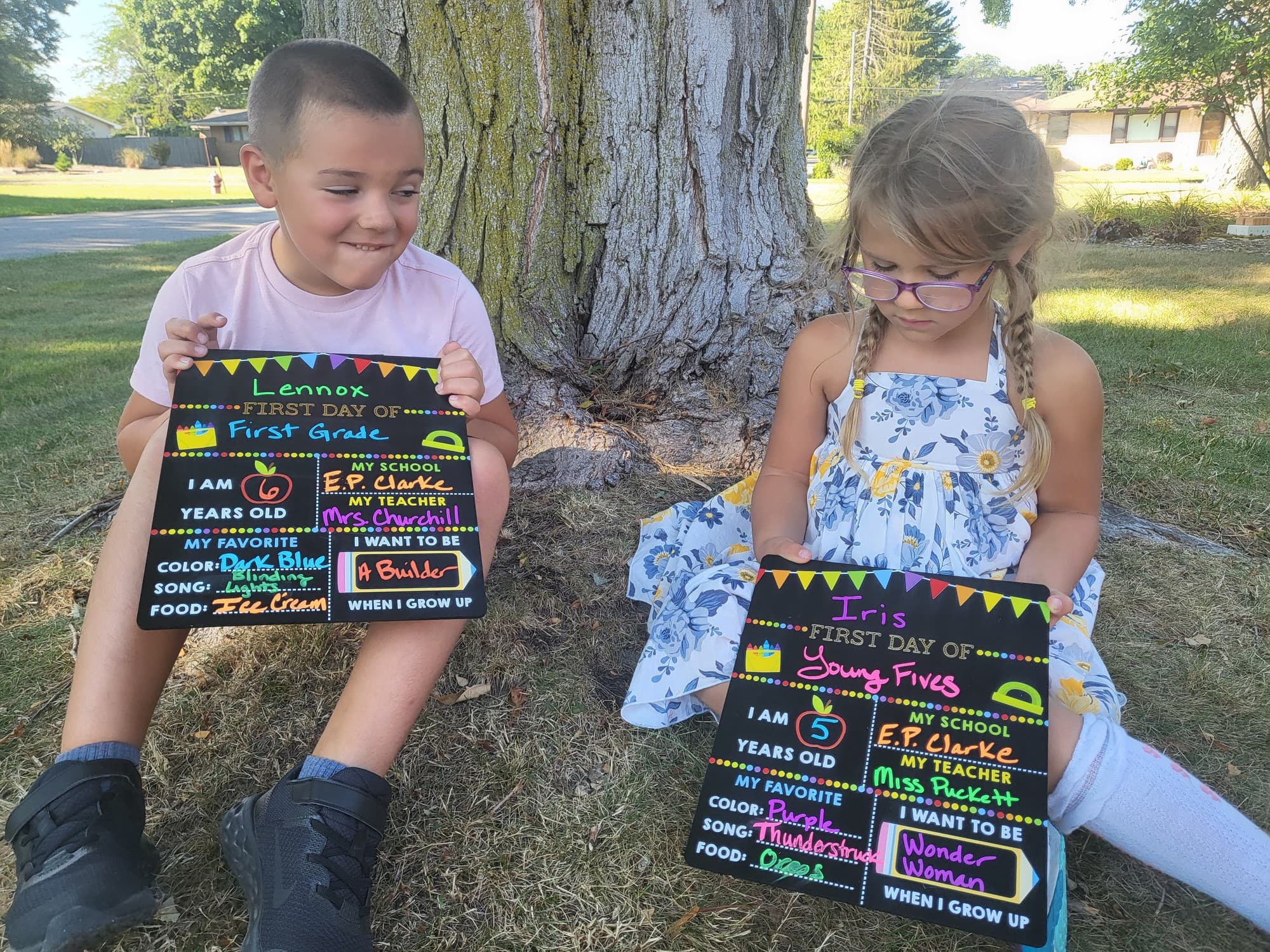 Lennox and Iris sitting under a tree holding first-day-of-school signs for first grade and young fives