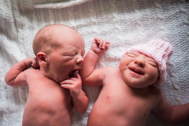 Newborn twins Lennox and Ella lying side by side on a white blanket, Ella smiling in a pink knit hat